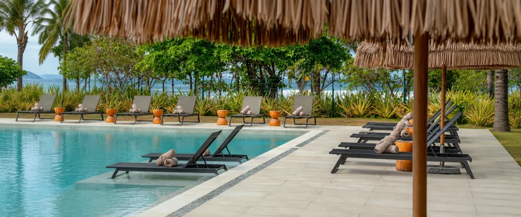 A clear resort swimming pool lined with dark lounge chairs, rolled towels, and thatched umbrellas, surrounded by lush tropical greenery and ocean views.